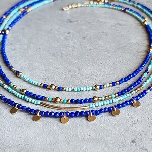May include: A layered necklace with blue and turquoise beads and gold accents. The necklace has a delicate gold bar pendant and small gold disc charms.