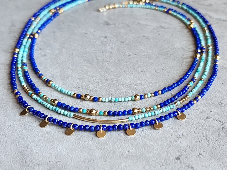 May include: A layered necklace with blue and turquoise beads and gold accents. The necklace has a delicate gold bar pendant and small gold disc charms.