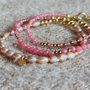 Puede incluir: Tres pulseras delicadas con piedras rosas, perlas y cuentas doradas. Las pulseras est&aacute;n superpuestas, creando un conjunto visualmente atractivo. Las perlas tienen un brillo suave y las piedras est&aacute;n facetadas.