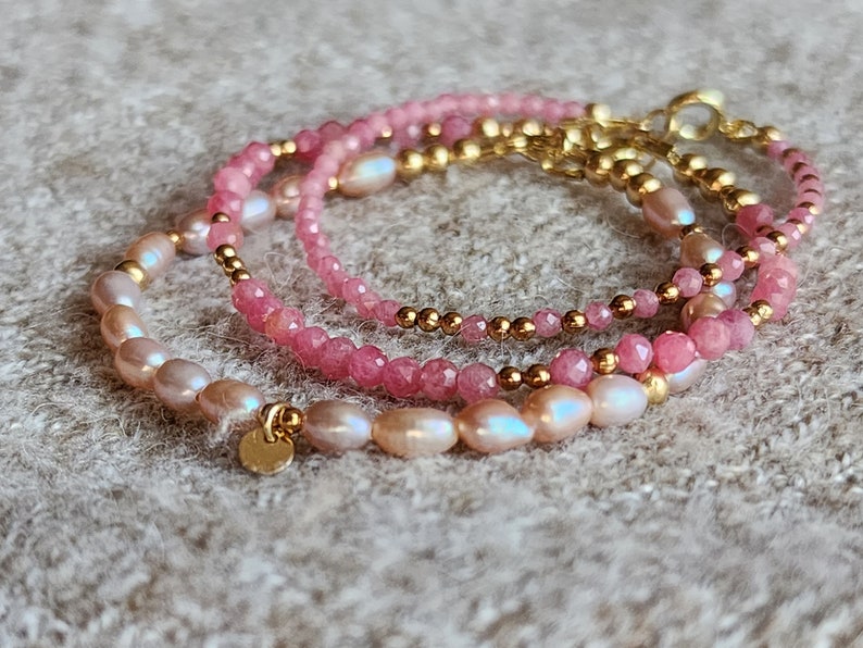 Puede incluir: Tres pulseras delicadas con piedras rosas, perlas y cuentas doradas. Las pulseras est&aacute;n superpuestas, creando un conjunto visualmente atractivo. Las perlas tienen un brillo suave y las piedras est&aacute;n facetadas.