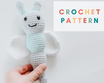 May include: A crocheted butterfly toy with white wings and a light blue and gray body. The toy has a smiling face with black eyes and a black mouth. The toy is being held in a hand.