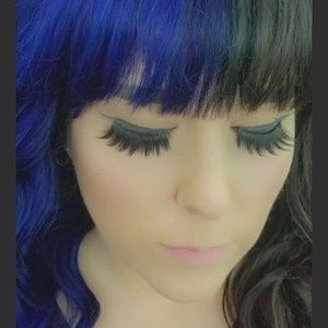May include: A person with long, dark hair parted with a vibrant blue section on the left. The person has long, dark eyelashes and is wearing a maroon top. The person's eyes are closed.