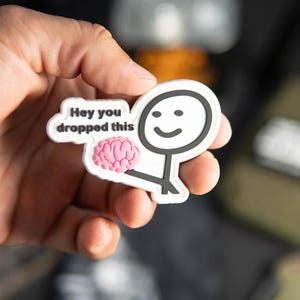 Könnte beinhalten: Ein weißer Gummi-Aufnäher mit einem schwarzen Strichmännchen, das ein rosa Gehirn hält. Eine Sprechblase neben der Figur enthält den schwarzen Text "Hey you dropped this". Der Aufnäher hat ein erhabenes, geformtes Design mit weißem Rand.