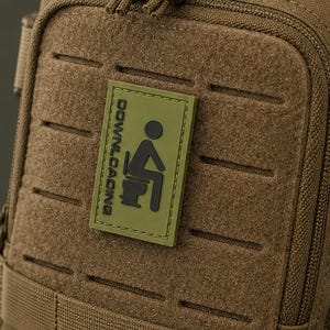 Könnte beinhalten: Ein beigefarbener taktischer Sling-Rucksack mit mehreren Reißverschlussfächern, verstellbaren Nylongurten und einem rechteckigen olivgrünen Aufnäher auf der Vorderseite. Der Aufnäher zeigt ein schwarzes Symbol einer Person auf einer Toilette mit dem Text DOWNLOADING.