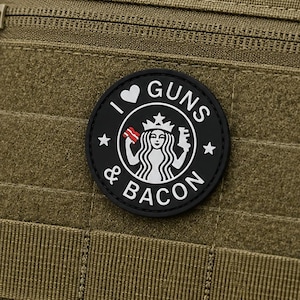 Puede incluir: Una mochila táctica de color verde oliva con un parche circular negro que presenta el texto blanco I HEART GUNS AND BACON. El parche muestra una figura central sosteniendo un trozo de tocino y un arma de fuego, rodeada por un borde y estrellas.
