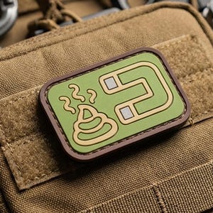 May include: A rectangular morale patch with a green background and a dark brown border attached to a tan tactical pouch. The patch features a graphic of a brown pile of excrement with steam lines next to a silver and tan horseshoe-shaped magnet.