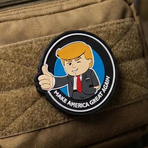 Puede incluir: Un parche circular con una ilustración de dibujos animados de una persona con cabello rubio, traje oscuro y corbata roja, haciendo un gesto de pulgar hacia arriba. El texto "MAKE AMERICA GREAT AGAIN" aparece en blanco en el borde negro.
