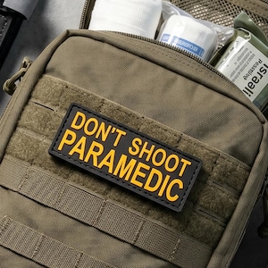 Può includere: Una borsa medica tattica verde oliva con una toppa nera e gialla con la scritta DON'T SHOOT PARAMEDIC. La borsa aperta contiene forniture mediche, accompagnate da forbici da trauma nere, un laccio emostatico, guanti in nitrile e una benda.