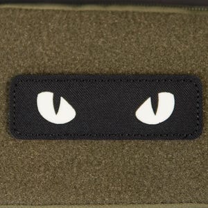 Op de afbeelding: Een olijfgroene rechthoekige etui met een ritssluiting. Een zwart label met witte oogvormen is in het midden aan de voorkant geplaatst. De etui heeft een lus en een ritstrekker.