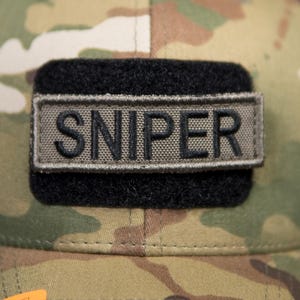 以下が含まれることがあります： 黒いメッシュの背面と湾曲したつばが付いた迷彩柄の野球帽。帽子には「SNIPER」の文字が刺繍された長方形のパッチが付いています。オレンジとグリーンの「MULTICAM」ステッカーがつばに貼られています。