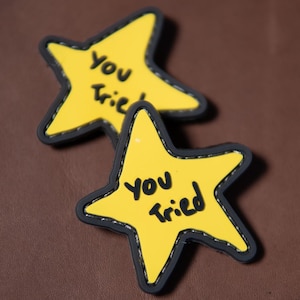May include: Two yellow star-shaped patches with black borders and the handwritten text "You Tried". The patches are on a brown surface. The stars have a stitched border.