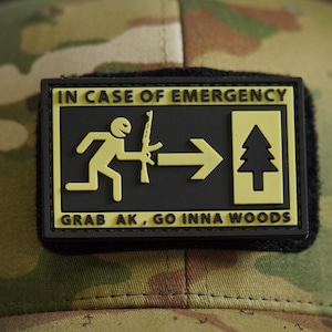 May include: A camouflage baseball cap featuring a rectangular black and yellow morale patch. The patch displays a stick figure holding a rifle, an arrow, and a pine tree, with the text IN CASE OF EMERGENCY GRAB AK, GO INNA WOODS.