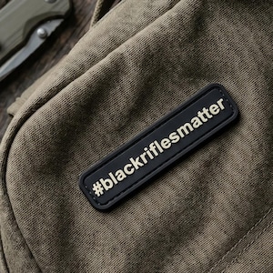 Puede incluir: Una bolsa de lona verde oliva con un parche rectangular negro que tiene el texto blanco #blackriflesmatter. Los objetos cercanos incluyen una linterna metálica, una navaja plegable verde oliva, un mosquetón negro con llaves y una cartera de cuero marrón.