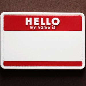 Peut inclure: Un badge d'identification blanc avec une bordure supérieure rouge. Le texte "HELLO my name is" est imprimé sur la bordure supérieure.