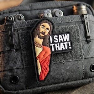 May include: A charcoal gray tactical pouch featuring a rubberized morale patch of a figure with a beard and red robe, accompanied by the white text "I SAW THAT!" on a black background. The pouch includes MOLLE webbing, metal clips, and a flashlight.