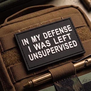 May include: A brown tactical pouch featuring a rectangular black morale patch with white text that reads IN MY DEFENSE I WAS LEFT UNSUPERVISED. The pouch includes a brass pen held in a fabric loop and a small green notebook tucked into a front pocket.