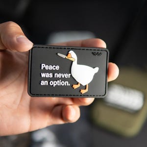 Op de afbeelding: Een rechthoekige zwarte patch met een witte gans die een zilverkleurig mes in de snavel houdt. De tekst "Peace was never an option." staat in witte letters aan de linkerkant. De gans heeft oranje poten en een oranje snavel.