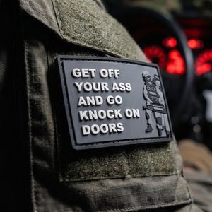 Può includere: Una toppa rettangolare nera con scritte bianche in rilievo che recitano GET OFF YOUR ASS AND GO KNOCK ON DOORS, con una grafica bianca di una figura tattica che impugna un fucile, fissata sulla manica color verde oliva di una divisa tattica.
