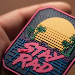 Könnte beinhalten: Ein rechteckiger Aufnäher mit Retro-Design. Der Aufnäher zeigt eine gelbe Sonne, Palmen und die Worte "STAY RAD" in Pink. Der Hintergrund ist schwarz mit einem blauen Gittermuster.