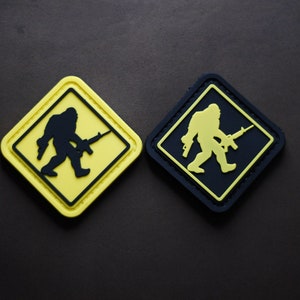 Könnte beinhalten: Zwei schwarze und gelbe Patches mit einer Silhouette eines Bigfoots, der ein Gewehr hält. Die Patches sind rautenförmig mit einem schwarzen Rand.