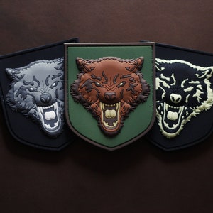 Könnte beinhalten: Drei gestickte Aufnäher mit einem Wolfskopf in verschiedenen Farbvarianten. Der Wolfskopf ist in einem Schwarz-, Braun- und Weißfarbschema gehalten. Die Aufnäher befinden sich auf einem braunen Hintergrund.