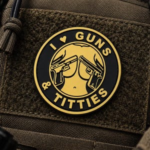 Puede incluir: Un parche moral circular negro y dorado sujeto a una bolsa táctica marrón. El parche presenta un gráfico de dos pistolas sobre un torso, con el texto I HEART GUNS AND TITTIES dispuesto en un borde circular alrededor del diseño central.