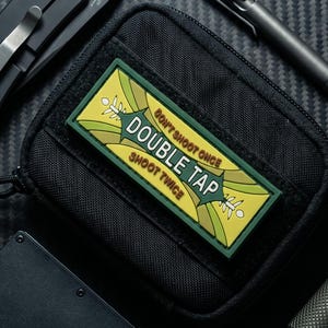 Puede incluir: Una bolsa táctica negra con un parche rectangular de diseño amarillo y verde. El parche muestra el texto "DON'T SHOOT ONCE DOUBLE TAP SHOOT TWICE". Alrededor de la bolsa hay una navaja plegable negra, un bolígrafo de metal y una linterna.