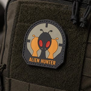 Peut inclure: Un sac tactique vert olive orné d'un écusson en caoutchouc noir, orange et rouge. L'écusson représente une silhouette extraterrestre stylisée dans un réticule de visée circulaire avec le texte ALIEN HUNTER imprimé en orange à la base.