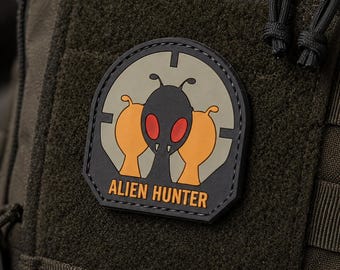 May include: An olive green tactical bag featuring a black, orange, and red rubber patch. The patch displays a stylized alien figure inside a circular target reticle with the text ALIEN HUNTER printed in orange letters at the bottom of the design.