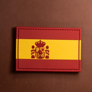 May include: A red and yellow fabric patch with the Spanish flag and coat of arms. The patch is stitched with a red thread around the edges.