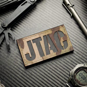 May include: A rectangular patch with the text JTAG in black block letters on a tan camouflage background, surrounded by a black multi-tool, a metallic pen, a digital wristwatch with a black nylon strap, and a black plastic buckle component.