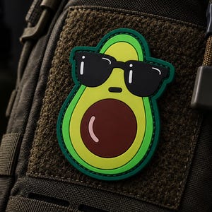 May include: A rubberized morale patch featuring a cartoon avocado wearing black sunglasses. The avocado has a bright yellow-green center, a dark brown pit with a white highlight, and a dark green border, attached to a dark olive green tactical bag.