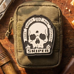 Könnte beinhalten: Eine olivgrüne taktische Tasche mit einem schwarzen und cremefarbenen Gummiaufnäher. Der Aufnäher zeigt ein Totenkopfmotiv mit dem Text "SHOOT THE FUCKERS" und "SNIPER" sowie ein Fadenkreuzsymbol. Die Tasche hat einen Reißverschluss.