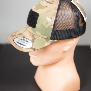 Può includere: Cappellino da baseball con motivo mimetico con retro in rete nera e toppa quadrata nera. Il cappellino ha una visiera curva e una chiusura a scatto. Il testo "SNAPBACK" è visibile sul davanti.