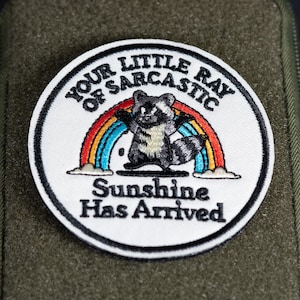 Peut inclure: Un écusson brodé circulaire représentant un raton laveur gris et blanc devant un arc-en-ciel coloré. Le texte indique : YOUR LITTLE RAY OF SARCASTIC Sunshine Has Arrived. L'écusson est fixé sur un étui rectangulaire en tissu vert olive.