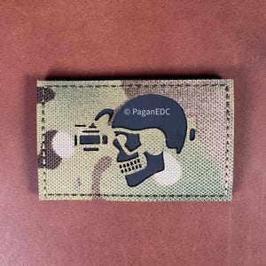 May include: A rectangular patch with a camouflage pattern background. A black skull graphic wearing a helmet and night vision device is centered on the patch. The text "© PaganEDC" is printed on the right side of the skull.