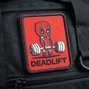 Peut inclure: Un sac de sport en tissu noir avec un écusson brodé rectangulaire. L'écusson présente un fond rouge avec un personnage de dessin animé soulevant une barre de musculation et le texte DEADLIFT en lettres capitales noires. Sangles en nylon noir.