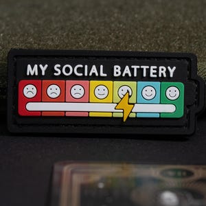 May include: A rectangular black patch featuring the white text MY SOCIAL BATTERY above a horizontal battery gauge. The gauge consists of seven colored squares with faces ranging from sad to happy, with a yellow lightning bolt icon over the fifth square.