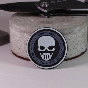 Könnte beinhalten: Ein schwarz-weißes Totenkopf-Patch mit dem Text "Don't Run You'll Only Die Tired".