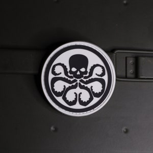 May include: A circular patch featuring a black graphic of a skull with six tentacles extending downwards, set against a white background with a thick black border. The design is centered on a dark olive green strap with a rectangular metal buckle.