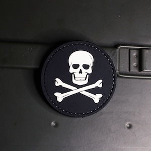 Può includere: Una toppa nera e bianca con un teschio e ossa incrociate su uno sfondo nero e cuciture bianche attorno al bordo.