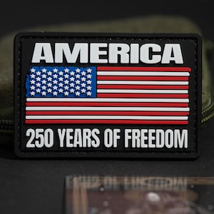 Peut inclure: Un écusson rectangulaire noir portant le texte blanc AMERICA en haut et 250 YEARS OF FREEDOM en bas. Au centre se trouve un drapeau américain stylisé avec un champ bleu parsemé d'étoiles blanches et des bandes horizontales rouges et blanches.
