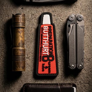 Könnte beinhalten: Eine Messing-Taschenlampe, ein rechteckiger rot-schwarzer Aufnäher mit dem Text PREPARATION BUTTHURT B cream & lube APPLY GENEROUSLY WHERE IT HURTS und ein graues Multifunktionswerkzeug. Die Gegenstände sind auf einer grauen Oberfläche angeordnet.