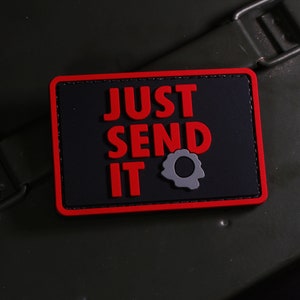 Puede incluir: Parche negro y rojo con el texto "JUST SEND IT" y un gráfico de agujero de bala gris.