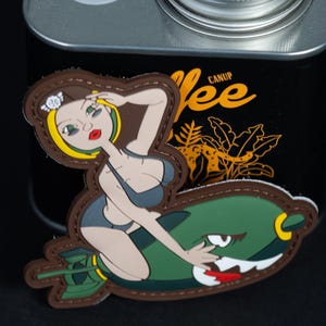 Könnte beinhalten: Ein gummiertes Aufnäher-Abzeichen mit einer Cartoon-Figur mit blonden Haaren und weißer Mütze, die einen dunkelgrauen Bikini trägt und auf einer grünen Bombe mit Haifischmaul-Grafik sitzt. Der Aufnäher hat einen braunen, genähten Rand.