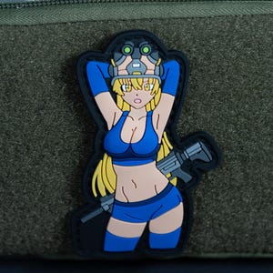 Könnte beinhalten: Ein Gummi-Aufnäher mit einer Comicfigur mit langem blondem Haar, die einen blauen Sport-BH, eine blaue Shorts und einen taktischen Helm mit grüner Nachtsichtbrille trägt. Die Figur hält ein graues Gewehr und ist vor einem dunklen Hintergrund.