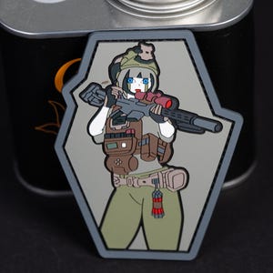 Könnte beinhalten: Ein sargförmiger PVC-Aufnäher mit einer Comicfigur mit blauen Augen und weißem Haar, die taktische Ausrüstung, einen Helm und eine braune Weste trägt. Die Figur hält ein schwarzes Gewehr mit rotem Zielfernrohr und hat einen roten Sprengkörper am Gürtel.