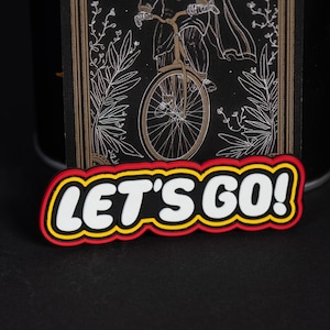 Puede incluir: Un parche rectangular negro con un dibujo lineal blanco de una persona en bicicleta rodeada de follaje. Debajo de la ilustración, un borde en capas rojo, amarillo y negro enmarca el texto blanco LET'S GO! en letras negritas.