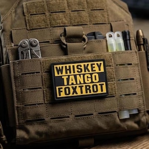 Può includere: Un gilet tattico portapiastre color marrone coyote con una toppa morale rettangolare nera e gialla con la scritta WHISKEY TANGO FOXTROT. Il gilet include cinghie MOLLE e tasche integrate contenenti un multiutensile, bastoncini luminosi e una penna.