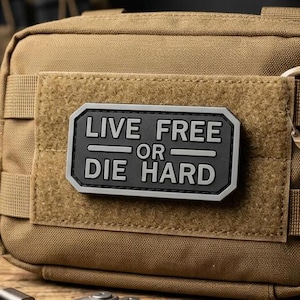 May include: A tan tactical utility pouch featuring a rectangular black patch with the white text "LIVE FREE OR DIE HARD" centered on the front. The pouch includes a small carabiner, a multi-tool, a flashlight, a charging cable, and a key ring.